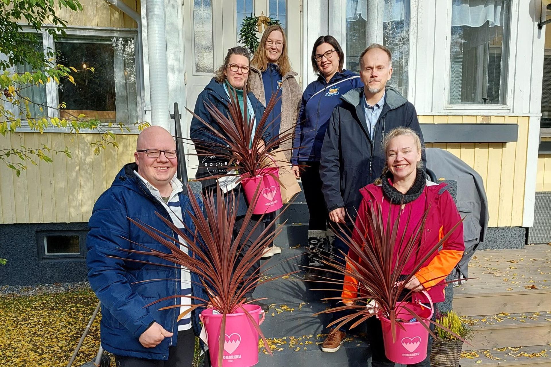 Asumiskokeilijat, Pomarkun kunnanjohtaja ja Palvelusihteeri-kirjastovirkailija, sekä hanketyöntekijät yhteiskuvassa Villa Rouheen ulkorappusilla. Käsissä asumiskokeilijoilla lahjaksi saadut pinkit ämpärit joissa pinkit kukat.