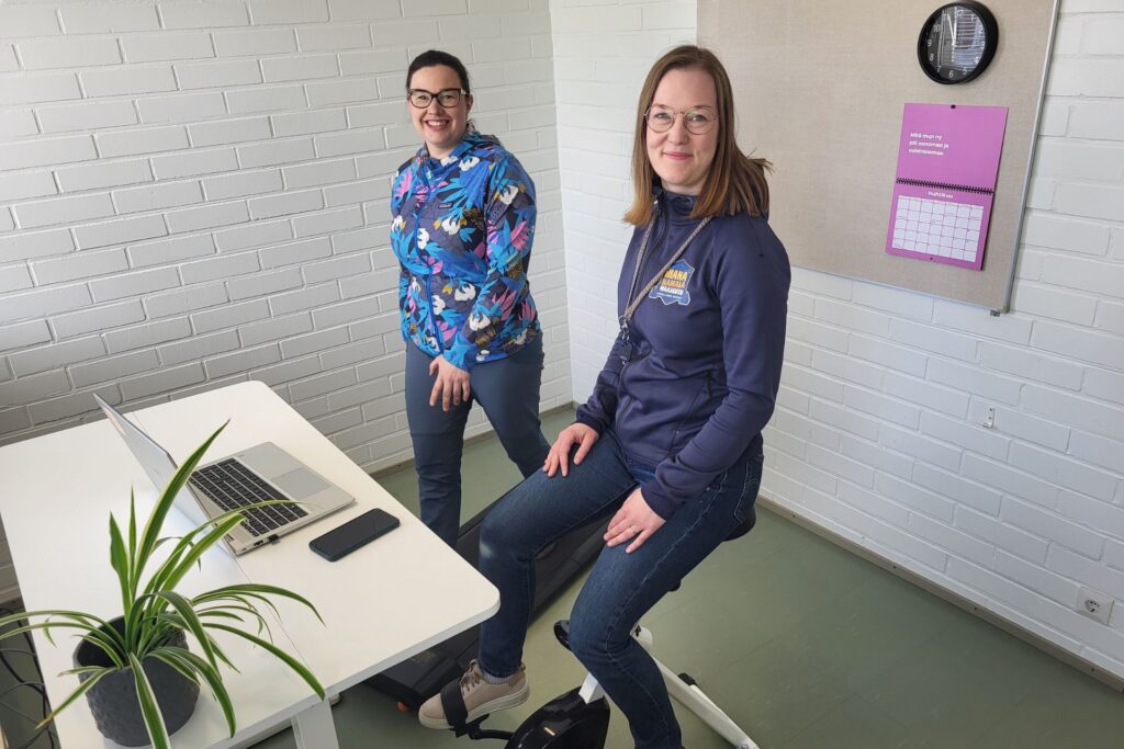 Merikarvian etätyötiloissa kaksi työntekijää, joista toinen kävelee kävelymatolla ja toinen polkee toimistopyörällä. Merikarvialla on panostettu hyvinvointiin ja tuotu etätyötiloihin liikuntavälineitä. 